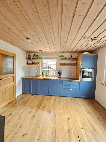 Een modern ingerichte keuken met blauwe kasten en een houten vloer. Natuurlijk licht valt door een raam, wat een aangename sfeer creëert.