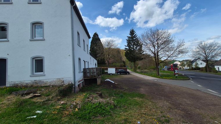Een witte gebouwen aan een straat, omringd door bomen en een groen gebied. De lucht is helder met enkele wolken.