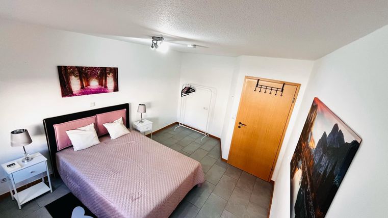 Een moderne slaapkamer met een tweepersoonsbed en roze beddengoed. Aan de muur hangen kunstwerken, en er is een nachtkastje en een kledingrek.
