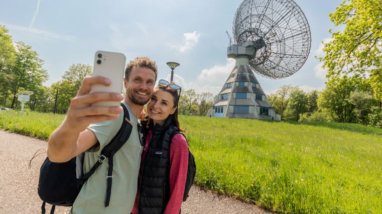 A couple takes a selfie in front of the Astropeiler Stockert radio telescope in a green landscape.