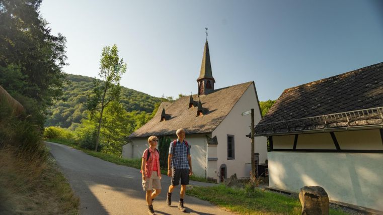 Zwei Personen spazieren an einem ruhigen Weg entlang. Im Hintergrund ist eine kleine Kirche und grüne Hügel zu sehen.