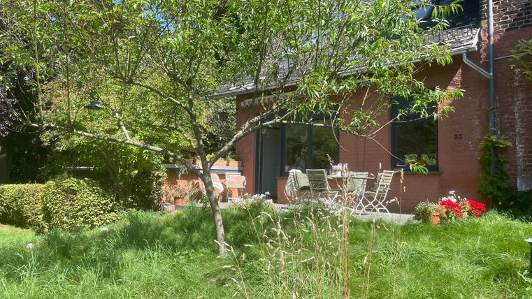 Un jardin confortable avec un arbre fruitier et un coin salon devant une maison rouge. La pelouse est verte et des plantes fleuries apportent de la couleur.