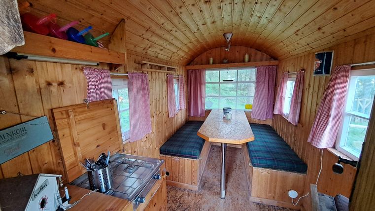 Ein gemütlicher Innenraum mit Holzverkleidung und einem großen Tisch in der Mitte. Die Fenster sind mit Vorhängen versehen und sorgen für viel Tageslicht.