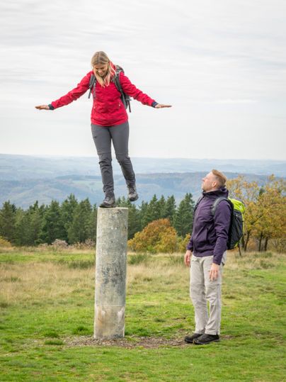 Eine Frau balanciert auf einem Betonpfosten, während ein Mann daneben steht und zuschaut. Im Hintergrund ist eine hügelige Landschaft zu sehen.