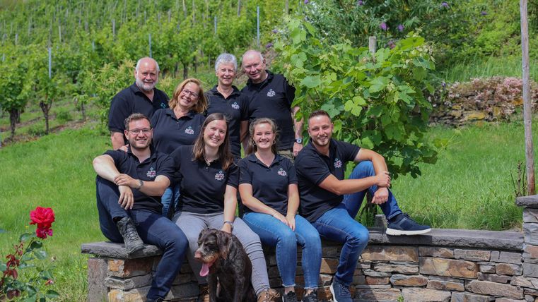 Een groep van acht personen zit glimlachend voor een wijngaard. Een hond ligt vooraan op de grond.