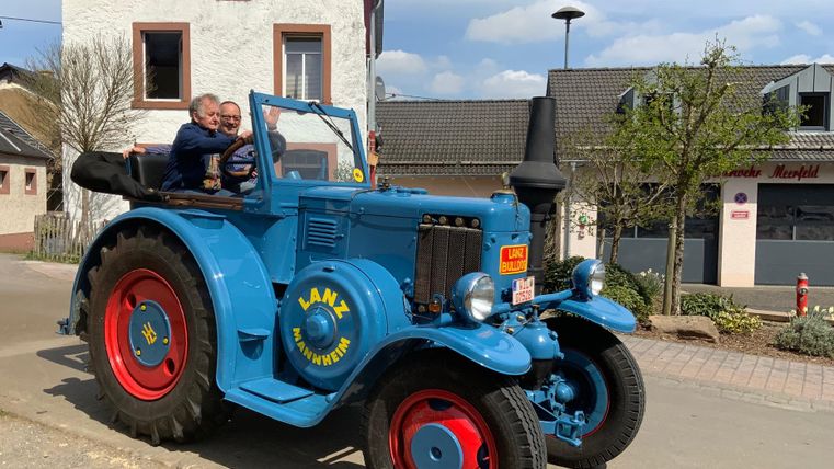 Ein blauer Oldtimer-Traktor steht auf einer Straße. Zwei Personen sitzen fröhlich auf dem Traktor.