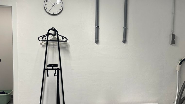 Ein moderner Raum mit grauen Fliesen und einer weißen Wand. Es gibt eine Uhr, einen Kleiderständer und eine Bank mit zwei Sitzkissen.