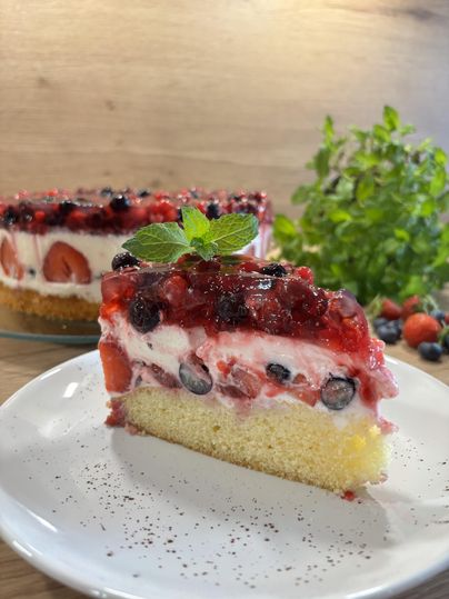 Een stuk bosvruchtentaart bestaande uit een laag bessen in gelei, daaronder een witte room en daaronder een cakebodem staat op een bord. Op de achtergrond de rest van de taart.