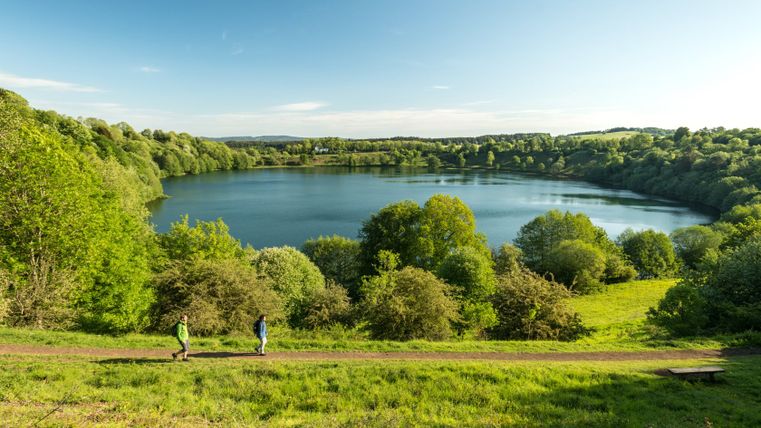 Twee wandelaars op een pad op het Weinfelder Maar in de Eifel, omgeven door een groen landschap en blauwe lucht.