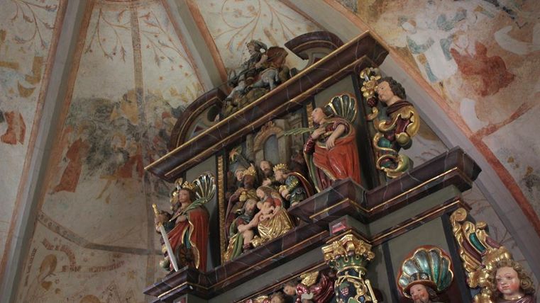 Ein kunstvoller Altar mit farbenfrohen Holzschnitzereien in einer Kirche. Die Wände sind mit alten Fresken verziert.