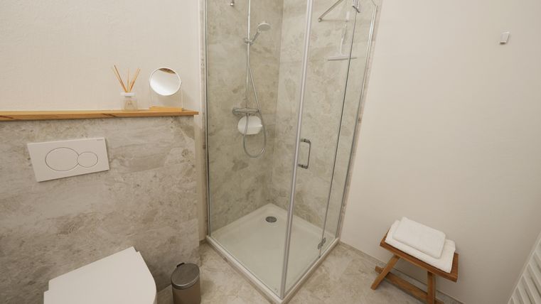 Une salle de bain moderne avec une cabine de douche, un WC et une étagère en bois. Des carreaux blancs et une ambiance claire et propre créent une atmosphère agréable.