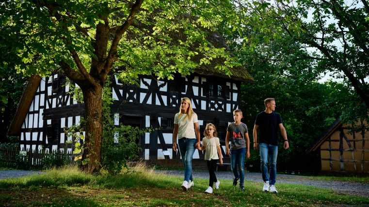 Eine Familie geht spazieren in der Natur vor einem hübschen, traditionellen Fachwerkhaus. Bäume umgeben die Szene und sorgen für eine entspannte Atmosphäre.