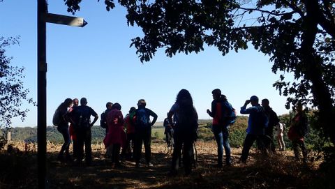 A group of hikers stands in front of a viewpoint. In the background, a clear sky and the landscape can be seen.