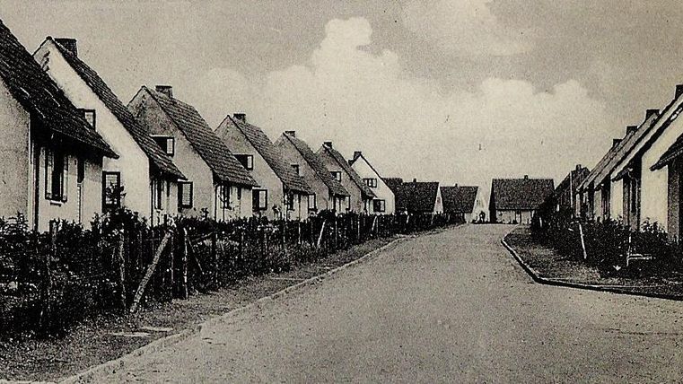 A quiet street with small, white houses on both sides. The sky is cloudy, and the surroundings seem rural.