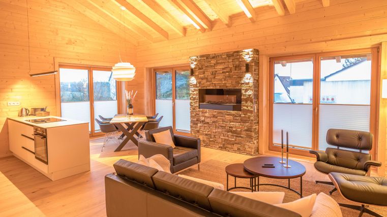 Ein modernes Wohnzimmer mit Holzwänden und großen Fenstern. Es gibt eine gemütliche Sitzecke, einen Esstisch und einen eleganten Kamin.