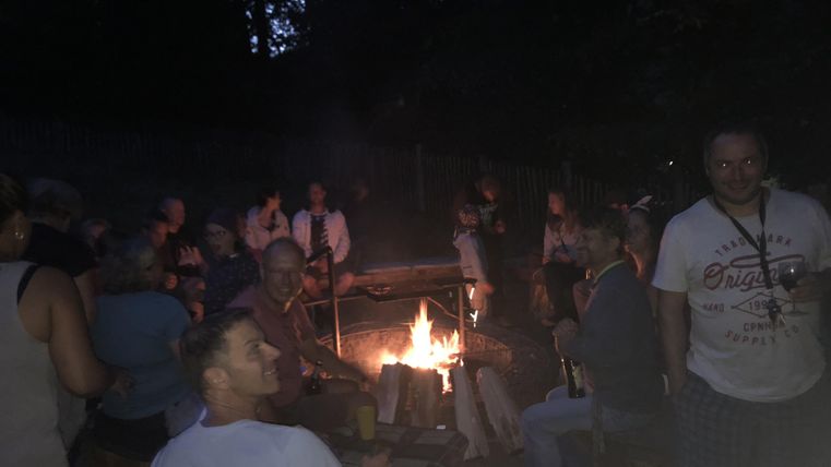 Un groupe de personnes est assis autour d'un feu de camp dans un environnement sombre. Une atmosphère chaleureuse et conviviale règne.