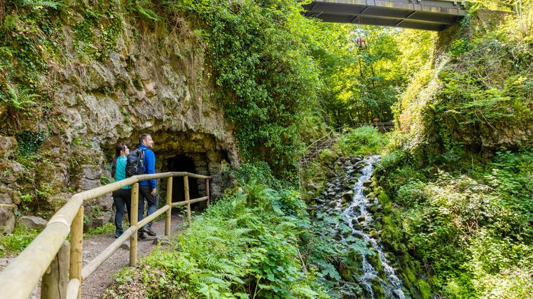 Twee mensen staan voor een grot in een groene, bosrijke omgeving met een kleine waterval en een brug erover.
