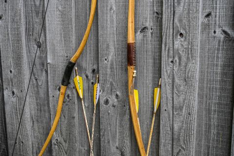 Zwei Bögen und Pfeile hängen an einer Holzwand. Die Pfeile haben gelbe Spitzen und sind sorgfältig angeordnet.