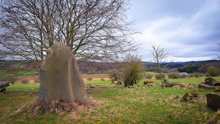 Ein großer Steinhaufen in einer grünen Wiese unter einem bewölkten Himmel. Im Hintergrund sind vereinzelte Bäume und Hügel zu sehen.