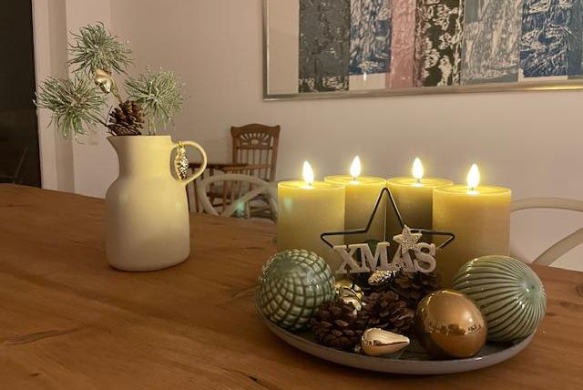 Ein gemütlicher Tisch mit einer Kerzenarrangement und Weihnachtsdekoration. Im Hintergrund steht eine Vase mit einem Tannenzweig.