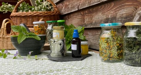 Ein Tisch mit verschiedenen Gläsern gefüllt mit Kräutern und natürlichen Ölen. Im Hintergrund sind Korb mit Pflanzen zu sehen.