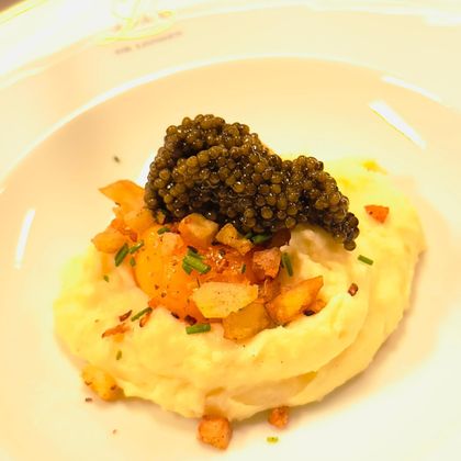 Een delicaat gerecht met romige aardappelpuree, bedekt met eigeel en zwarte kaviaar. Daarop liggen krokant gebakken stukjes van appel en bieslook.