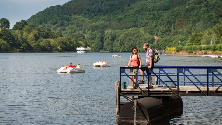 Een echtpaar staat op een steiger bij het meer in Einruhr, omringd door waterfietsen en een plezierboot. Op de achtergrond zijn beboste heuvels te zien.