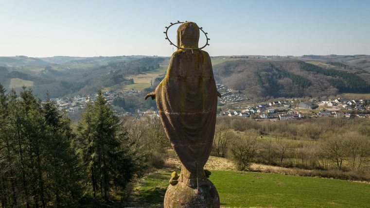 Eine große Statue einer Madonna steht auf einer Säule und blickt über die Landschaft. Im Hintergrund sind sanfte Hügel und ein kleines Dorf zu sehen.