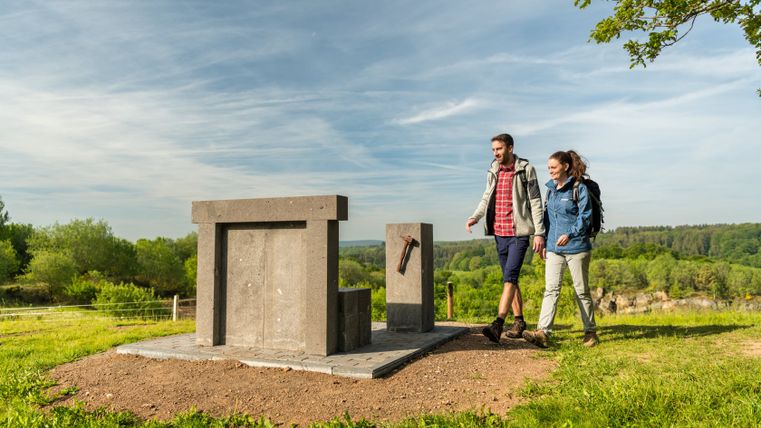 Zwei Wanderer gehen an einer Steinskulptur vorbei, die wie ein Thron aussieht, auf einem grünen Hügel mit bewaldetem Hintergrund.