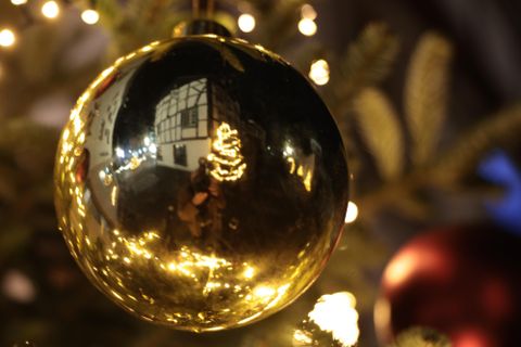 A shiny, golden Christmas ornament hangs on a tree. The ball reflects a festively decorated room with lights.