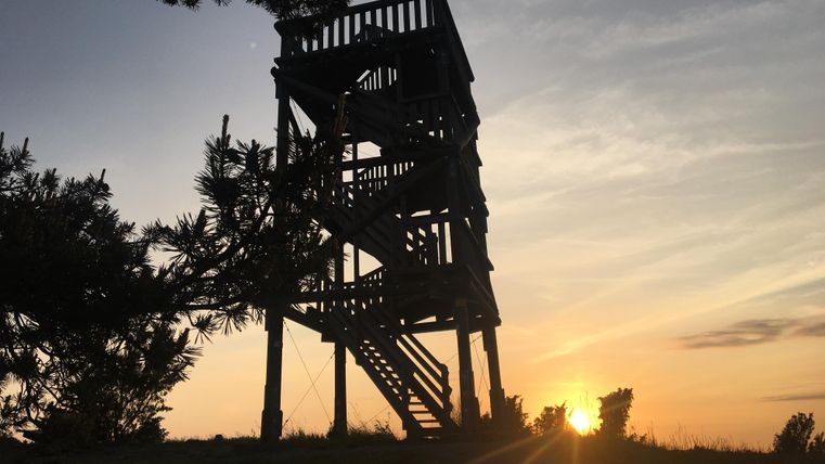 Ein Aussichtsturm steht silhouettiert gegen den Sonnenuntergang. Der Himmel ist orange und rosa gefärbt, während das Licht mild über die Landschaft strahlt.
