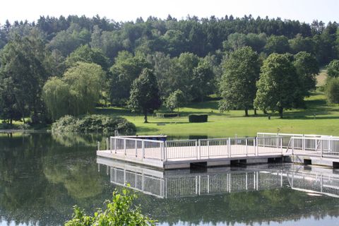 A calm lake with a wooden platform in the foreground. In the background, green trees and gentle hills can be seen.