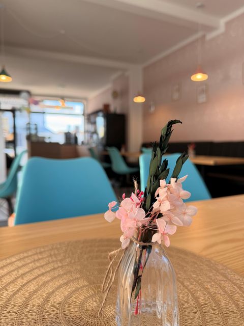 Een gezellig café met blauwe stoelen en warme verlichting. Op de tafel staat een kleine glazen vaas met roze bloemen.