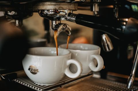 Twee witte koffiekopjes staan onder een espressomachine. Verse espresso stroomt uit de tuit.