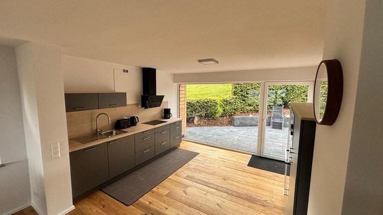 Een moderne keuken met grijze kasten en een houten vloer. Grote ramen bieden uitzicht op de buitenruimte met tuin.