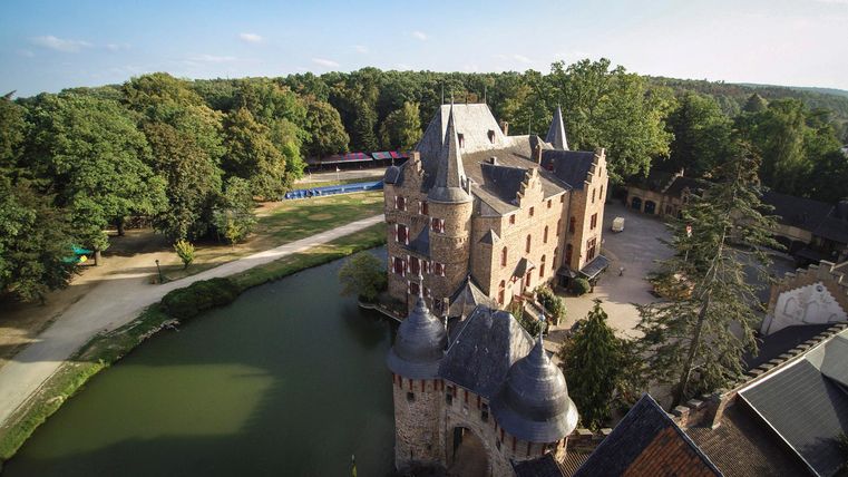 Eine beeindruckende Burg mit Türmen, umgeben von einem Wassergraben. Das grüne Umland und die Bäume schaffen eine malerische Atmosphäre.