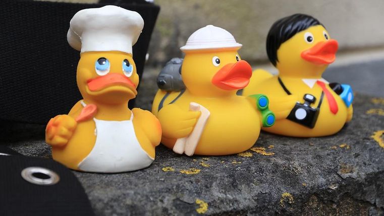 Drei Gummienten nebeneinander auf einer Mauer in unterschiedlicher Verkleidung - eine Ente als Koch, eine als Entdecker und eine als Reporter.