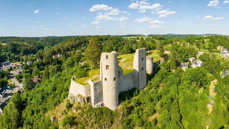 Eine alte Burgruine auf einem Hügel, umgeben von grünen Wäldern. Im Hintergrund sind kleine Häuser und ein klarer Himmel zu sehen.