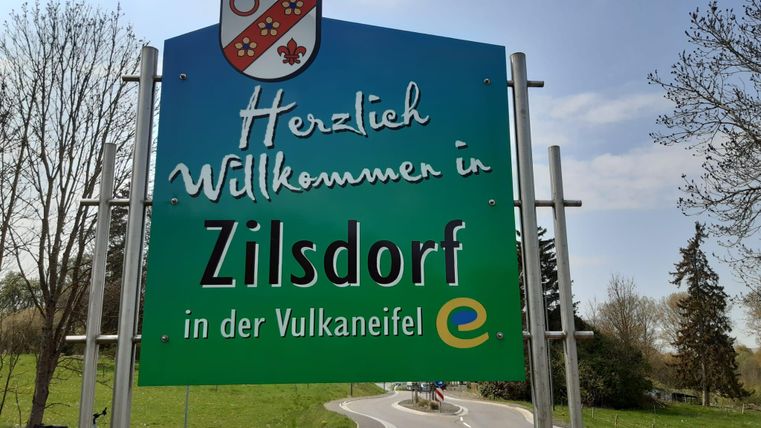 A welcome sign for Zilsdorf in the Vulkaneifel. The surroundings are green and provide a rural backdrop.