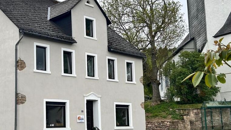 Ein weißes Haus mit mehreren Fenstern und einer dunklen Schieferdach. Neben dem Gebäude steht ein großer Baum in einer ruhigen Straße.
