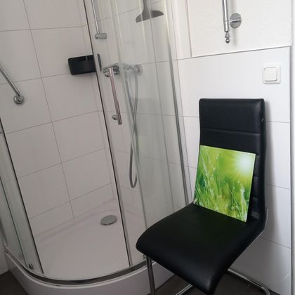 Een moderne douche staat in een heldere badkamer. Op een zwarte stoel ligt een afbeelding met groene plantenmotieven.