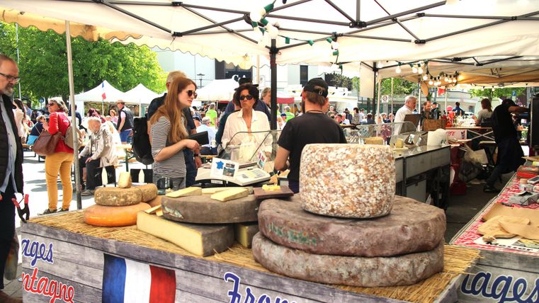 A market stall with various types of cheese and happy customers. In the background, there are additional stalls and visitors to be seen.