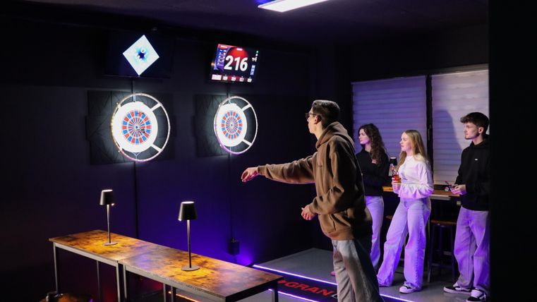 Een persoon speelt darts in een moderne ruimte met digitale dartborden. Op de achtergrond staan drie andere personen en bekijken ze het spel.