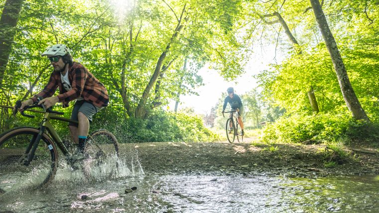 Two cyclists on gravel bikes ride through a large puddle in the middle of the forest. The sun shines through the trees.