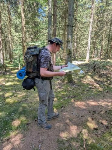A man stands in the forest and studies a map. He is wearing a backpack and is intently looking at the plan.