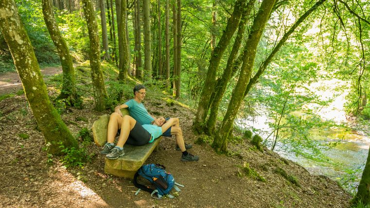 Zwei Personen sitzen auf einer Holzbank im schattigen Wald, mit Blick auf einen ruhig fließenden, sonnenbeschienenen Fluss. Sie tragen sportliche Outdoor-Kleidung, ein Rucksack liegt neben der Bank. Umgeben von hohen Bäumen genießen sie eine ruhige Pause in der Natur.