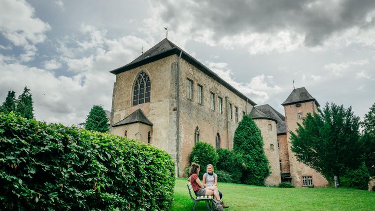 Een oude kerk omringd door groenvelden en bomen. Twee personen zitten ontspannen op de voorgrond op een gazon.