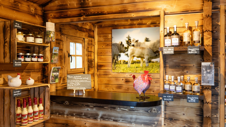 Ein gemütlicher Verkaufsraum aus Holz mit Regalen voller Lebensmittel und Getränken. An der Wand hängt ein Bild von Kühen auf einer Wiese.
