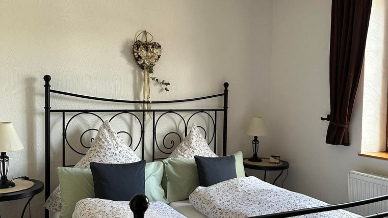 Een gezellige slaapkamer met een iron bed en decoratieve kussens. Er zijn twee nachtkastjes met lampen en een groot raam met gordijnen.