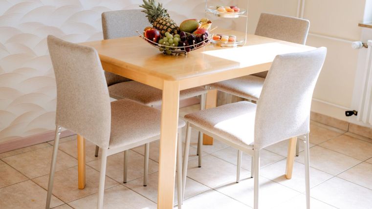 A bright dining area with a wooden table and four chair-like armchairs. On the table, there is a fruit basket with various fruits.